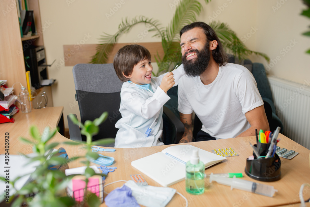 Obraz premium Little boy playing pretends like doctor examining a man in comfortabe medical office. Healthcare, childhood, medicine, protection and happiness concept. Having fun, laughting while examining a