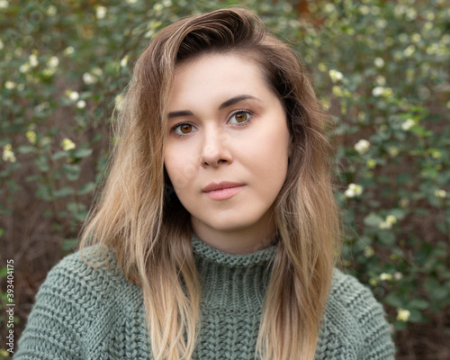 Young beautiful 27 years old woman in a sweater, standing outdoors against a background of greenery, sincerely and expressively looking into the camera. Close-up photo