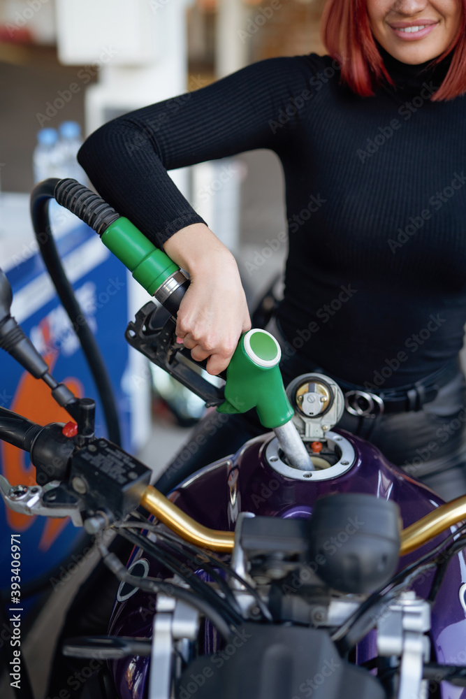 Obraz premium A slim woman fueling her modern motorcycle in refueling with gun. A refueling in sunny day. Custom urban bike.