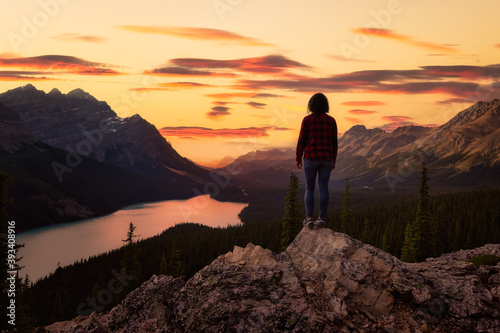 Adventurous girl standing on the edge of a cliff overlooking the beautiful Canadian Rockies and Peyto Lake. Dramatic, vibrant summer sunset. Taken in Banff National Park, Alberta, Canada.