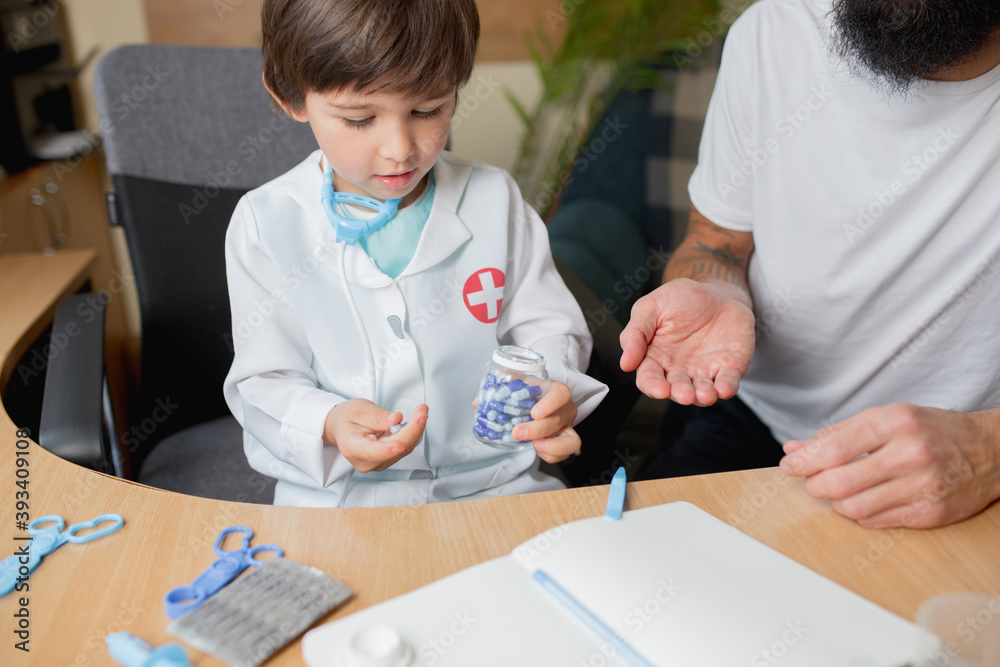 Little boy playing pretends like doctor examining a man in comfortabe medical office. Healthcare, childhood, medicine, protection and happiness concept. Having fun, laughting while giving pills Little boy playing pretends like doctor examining a man in comfortabe medical office. Healthcare, childhood, medicine, protection and happiness concept. Having fun, laughting while giving pills