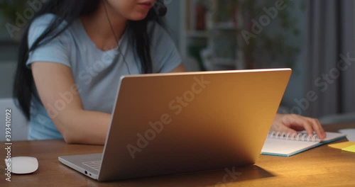 Close up portrait of young Asian female sitting at table and speaking on headset while working on laptop. Joyful beautiful woman having call in garniture and typing on computer indoor. Work concept