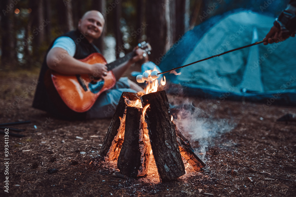 Marshmallow on a stick toasting on bonfire in summer forest in background of hairless mature guy he plays guitar.