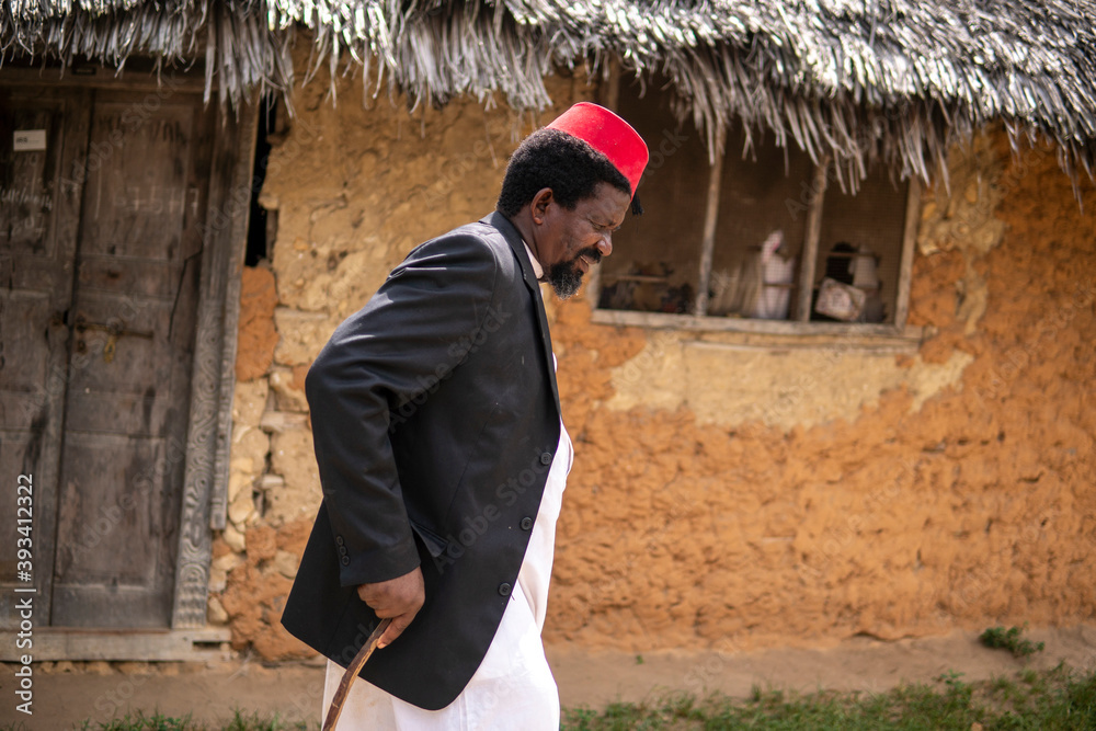 An African Older Man in Red Muslim Taqiyyah Fez Hat And Blazer Walking with a stick for limping people Near the Basic Hut with Thatched roof in Small Remote Village in Tanzania, Pemba island An African Older Man in Red Muslim Taqiyyah Fez Hat And Blazer Walking with a stick for limping people Near the Basic Hut with Thatched roof in Small Remote Village in Tanzania, Pemba island