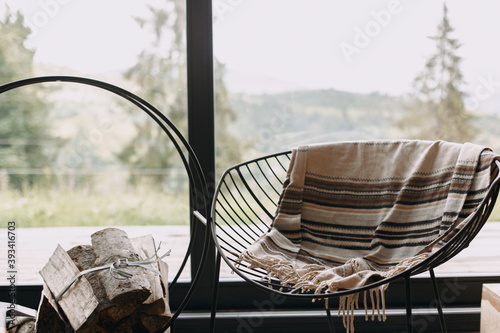Fototapeta Modern chair with cozy blanket and firewood on metal stand at window view on mou