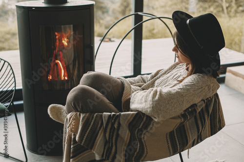 Fotografia Stylish woman in knitted sweater and hat warming up at modern black fireplace