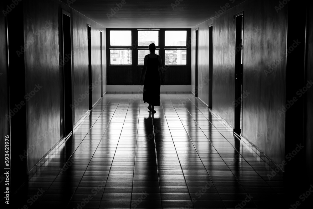Figura feminina caminhando em um corredor, iluminada, em contraluz, por uma janela. Imagem em preto e branco.