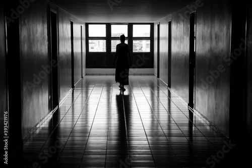 Figura feminina caminhando em um corredor  iluminada  em contraluz  por uma janela. Imagem em preto e branco.