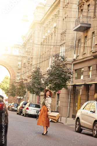 Full length of a stylish happy woman wearing grey coat, hat and high heel shoes standing with shopping bags on city street and smiling at camera