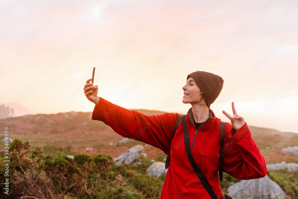 Attractive young woman in red clothes taking pictures at sunset in the field. He wears red clothes and a woolen hat. makes the symbol of victory. Attractive young woman in red clothes taking pictures at sunset in the field. He wears red clothes and a woolen hat. makes the symbol of victory.