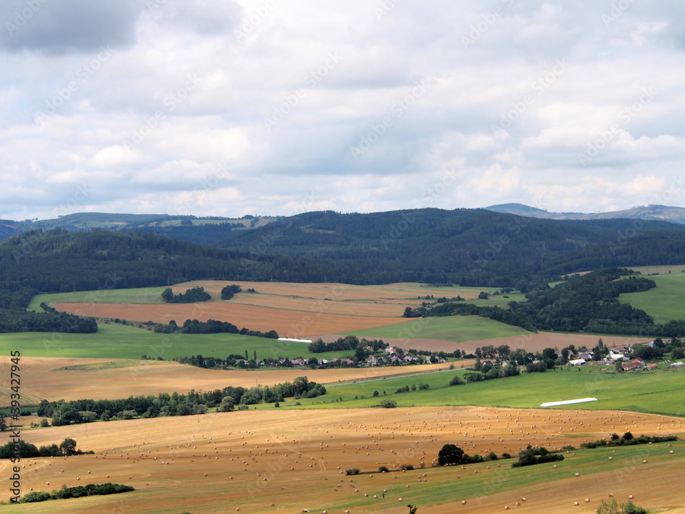 view of small town in fields, agricultural landscape, round bales of straw in fields, forests and hills in background, summer