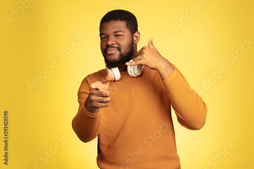 Pointing, choosing. Young african-american man's portrait isolated on yellow studio background, facial expression. Beautiful male model with headphones, copyspace. Concept of human emotions, sales, ad