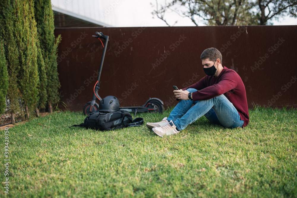 Fototapeta premium Young man using a mobile phone with an electric skate sitting on the grass.