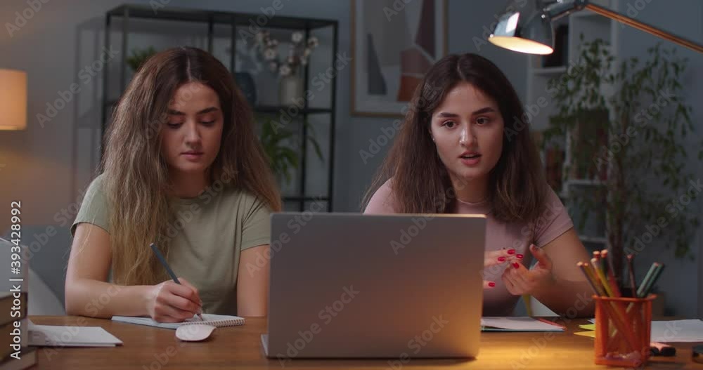 Portrait of two Caucasian twin sisters sitting in room and studying while having online lesson on laptop Pretty female writing in notebook while sister talking on video on computer distance education