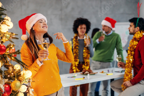 Photography Young smiling cheerful businesswoman with santa's hat on head holding champagne, standing next to christmas tree and celebrating new year's eve