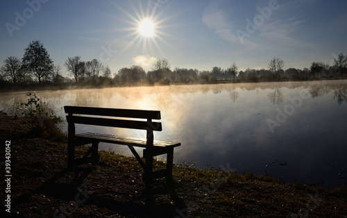 An old wooden bench stands by a lake at sunrise and rising mist in autumn
