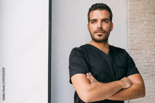 Male dentist in black medical scrubs looking away while standing with arms crossed against wall photo