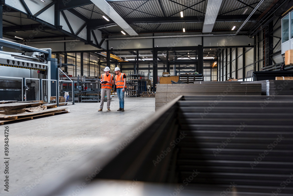 Two men wearing hard hats and safety vests talking on factory shop floor
