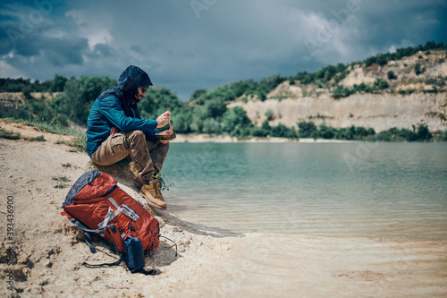 Young handsome bearded man sitting on a lake shore on a camping trip and eating his lunch. © chika_milan