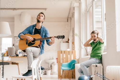 Horrified son covering his ears with father playing guitar at home photo