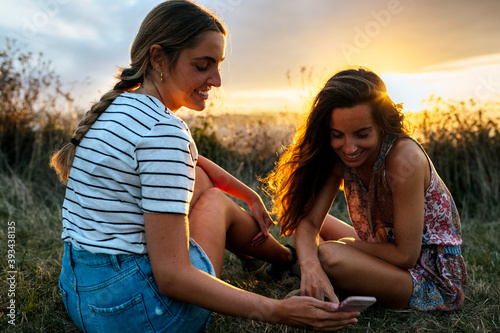 Smiling young female friends looking at smart phone while sitting on field during sunset photo