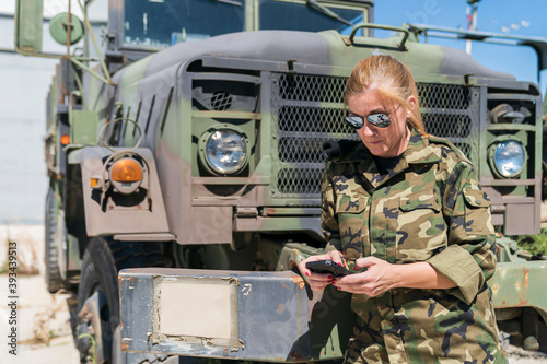 Mature blond female army soldier text messaging through smart phone while standing against truck at base on sunny day photo