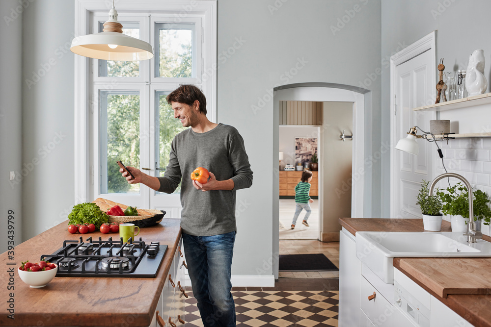 Smiling man using smartphone and holding bell pepper in kitchen Smiling man using smartphone and holding bell pepper in kitchen