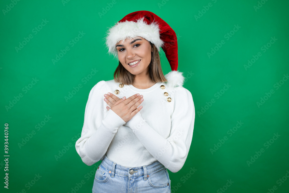 Young beautiful woman wearing a Santa hat and white sweater over green background smiling with her hands on her chest and grateful gesture on her face. Young beautiful woman wearing a Santa hat and white sweater over green background smiling with her hands on her chest and grateful gesture on her face.