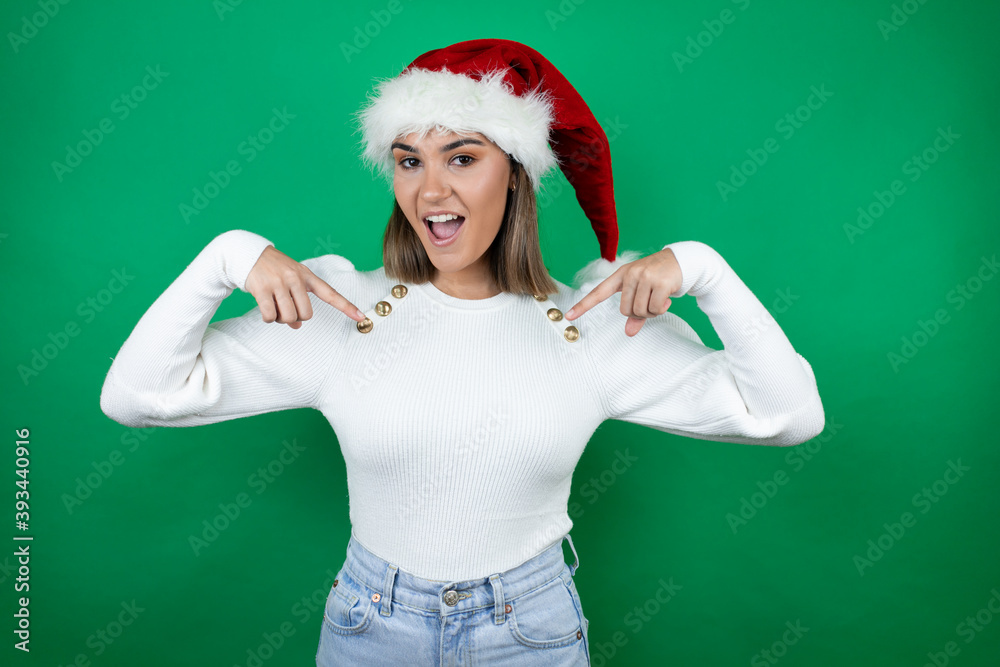 Young beautiful woman wearing a Santa hat and white sweater over green background looking confident with smile on face, pointing oneself with fingers proud and happy. Young beautiful woman wearing a Santa hat and white sweater over green background looking confident with smile on face, pointing oneself with fingers proud and happy.