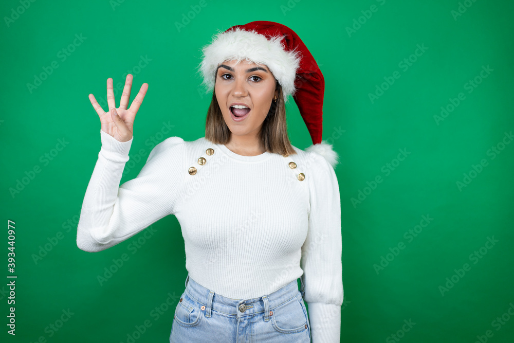 Young beautiful woman wearing a Santa hat and white sweater over green background showing and pointing up with fingers number four while smiling confident and happy Young beautiful woman wearing a Santa hat and white sweater over green background showing and pointing up with fingers number four while smiling confident and happy