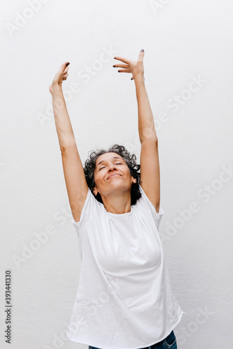 Mature woman standing with eyes closed and arms raised against white wall