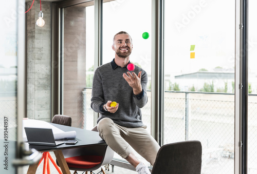 Smiling young businessman sitting at the window juggling with balls photo