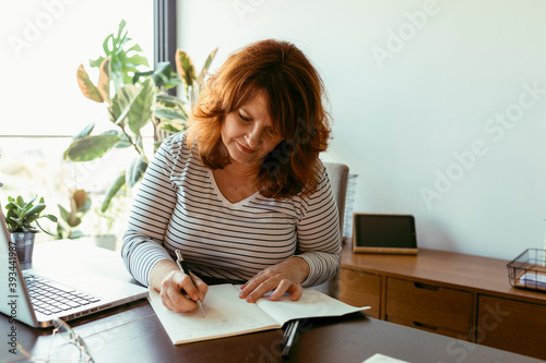 Woman writing in boo while using laptop on table at home