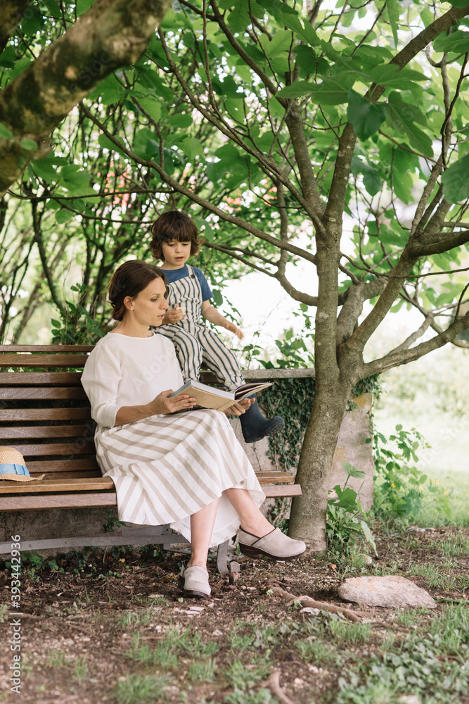 Beautiful woman and boy reading book while sitting on bench at backyard Beautiful woman and boy reading book while sitting on bench at backyard