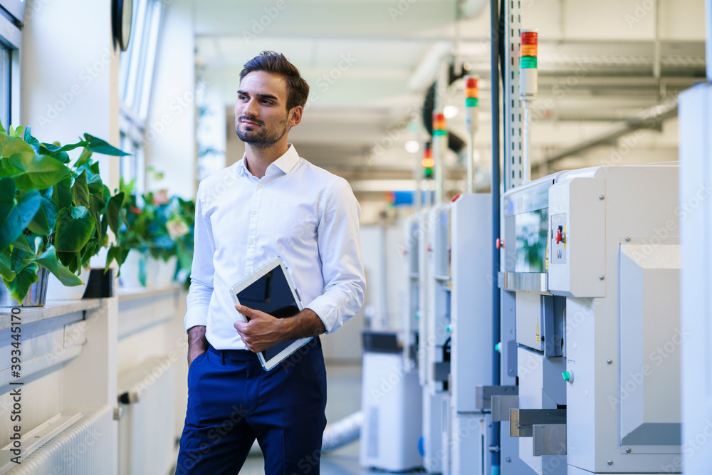Young businessman holding digital tablet while looking away and contemplating at illuminated factory
