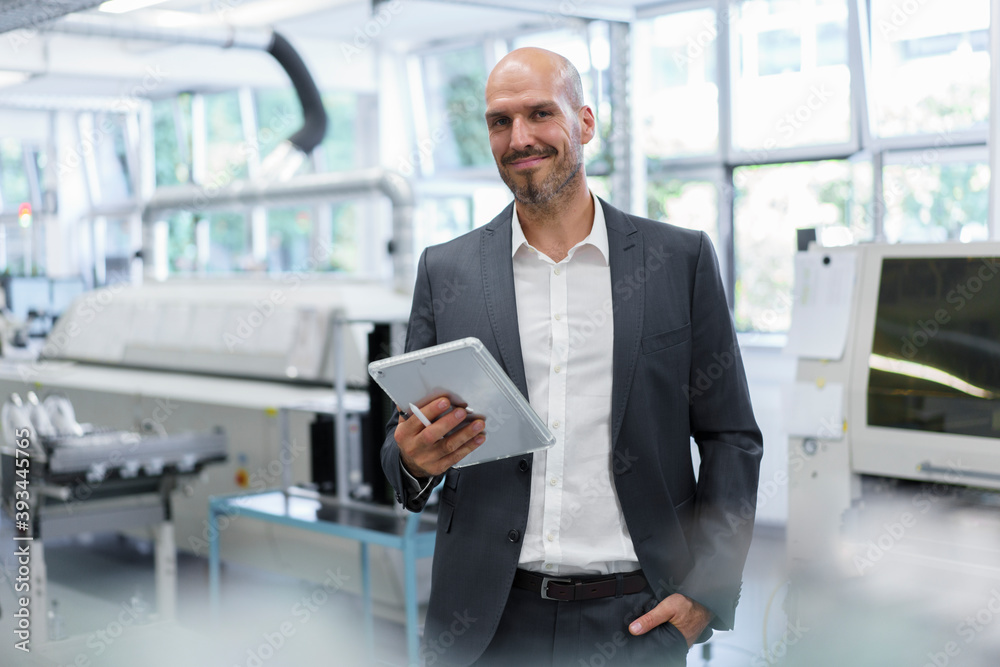 Smiling confident businessman holding digital tablet while standing with hand in pocket at bright factory Smiling confident businessman holding digital tablet while standing with hand in pocket at bright factory