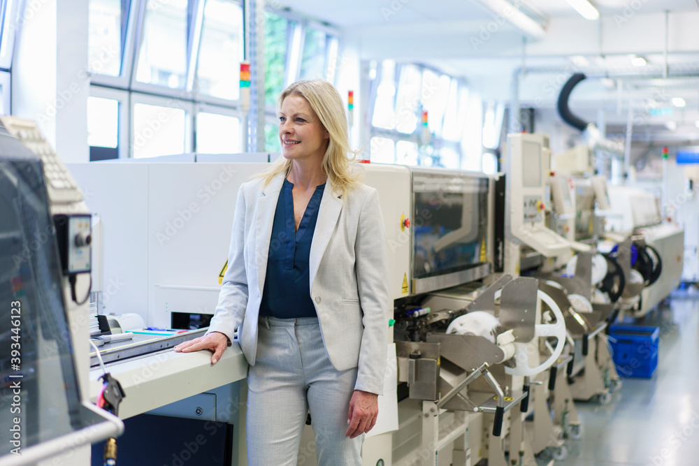 Thoughtful mature blond businesswoman standing by machinery while looking away at illuminated industry Thoughtful mature blond businesswoman standing by machinery while looking away at illuminated industry