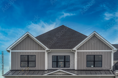 Upscale American house façade with vertical beige vinyl siding white accents, black framed aluminum windows metal roof cover over the porch, double gable roof with dreamy blue sky in the USA