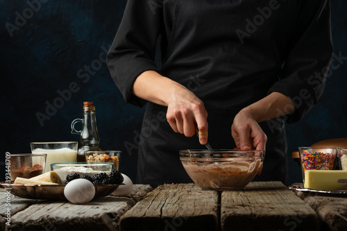 The professional chef kneads dough for preparing waffles on rustic wooden table with ingredients on dark blue background. Backstage of cooking sweet dessert for breakfast. Frozen motion. Good recipe.