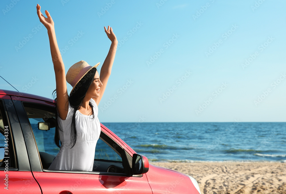 Fototapeta premium Happy young woman leaning out of car window on beach, space for text. Summer trip