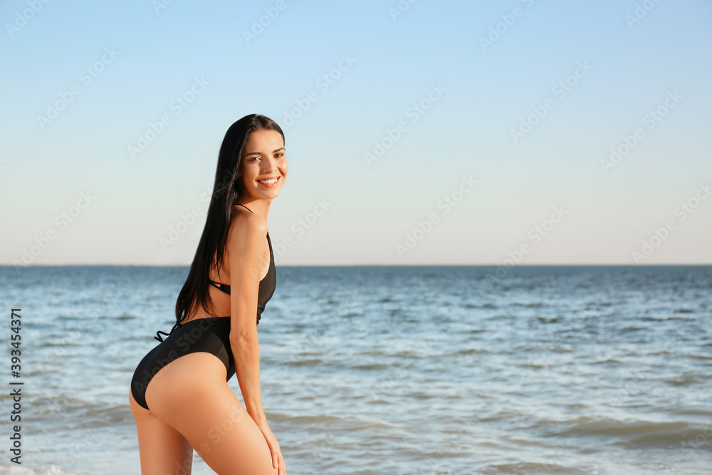 Beautiful young woman in black stylish swimsuit on beach. Space for text