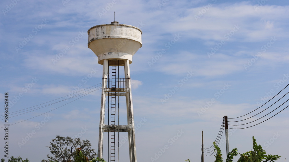 White concrete water tank on the tower. Water storage tanks for communities or establishments. Below are electric cables and green trees. On the sky background there are white clouds with a copy space