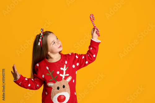 Cute little girl in Christmas sweater and festive headband holding candy cane on yellow background, space for text