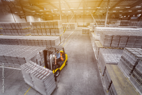 A forklift truck drives through a huge warehouse with stacks of vacuum-packed water or beer packages on pallets. View from above. Selective focus