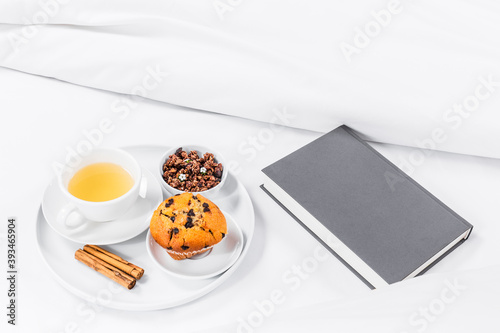 Breakfast tray on the bed, with tea, muffin, cinnamon and granola, white sheets and grey book. White background. Copy Space.