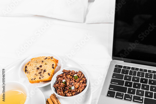 Breakfast tray on the bed, with tea, muffin, cinnamon and granola, white sheets, grey laptop. White background, copy space.