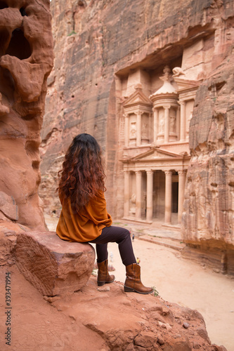 A girl in an orange shirt and cowboy boots sits on a stone and looks at the ancient city of Petra