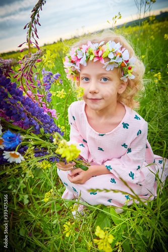 Portrait of pretty young small blonde girl with curly hair in pink dress in field with grass and flowers in summer day and cloudy sky background