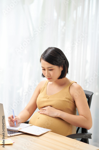 Pregnant freelancer touching belly near laptop, writing on diary on table