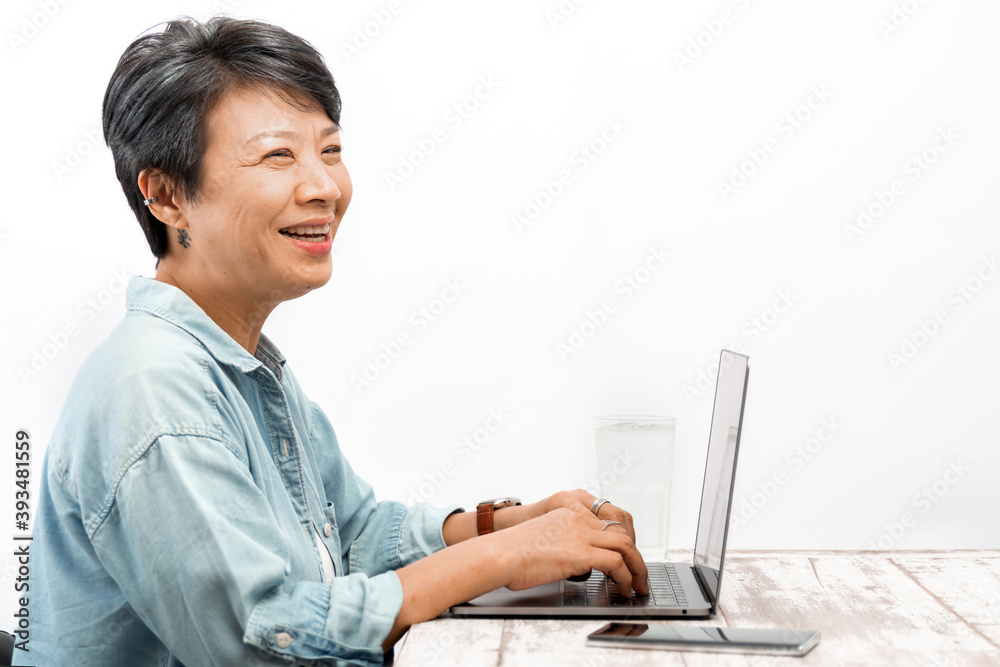 Senior businesswoman working on a laptop in office. Asian mature female with grey hair smiling at a laptop computer while sitting at her desk. Isolated white background. Active Elderly concept Senior businesswoman working on a laptop in office. Asian mature female with grey hair smiling at a laptop computer while sitting at her desk. Isolated white background. Active Elderly concept
